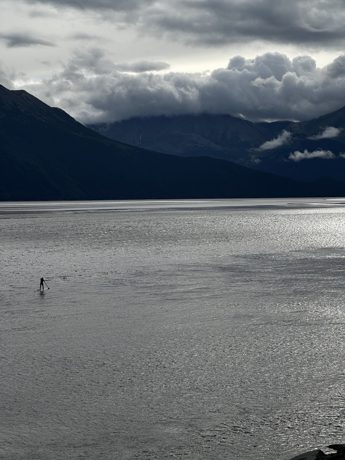 Paddleboarder silhouetted on Turnagain Arm during bore tide, storm clouds over Chugach Mountains
